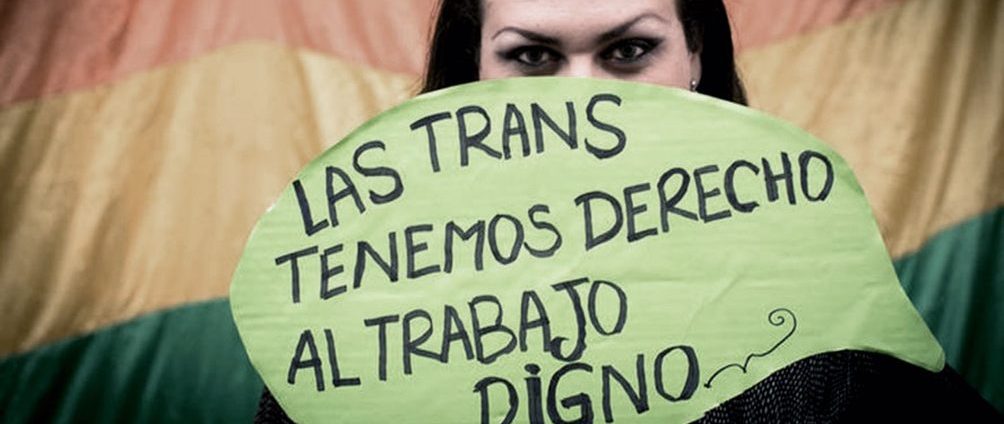 Una mujer sostiene un cartel, que le cubre la mitad de su cara, en el que está escrito el siguiente texto: "Las trans tenemos derecho al trabajo digno." Detrás de ella, hay una bandera del Orgullo LGTB+